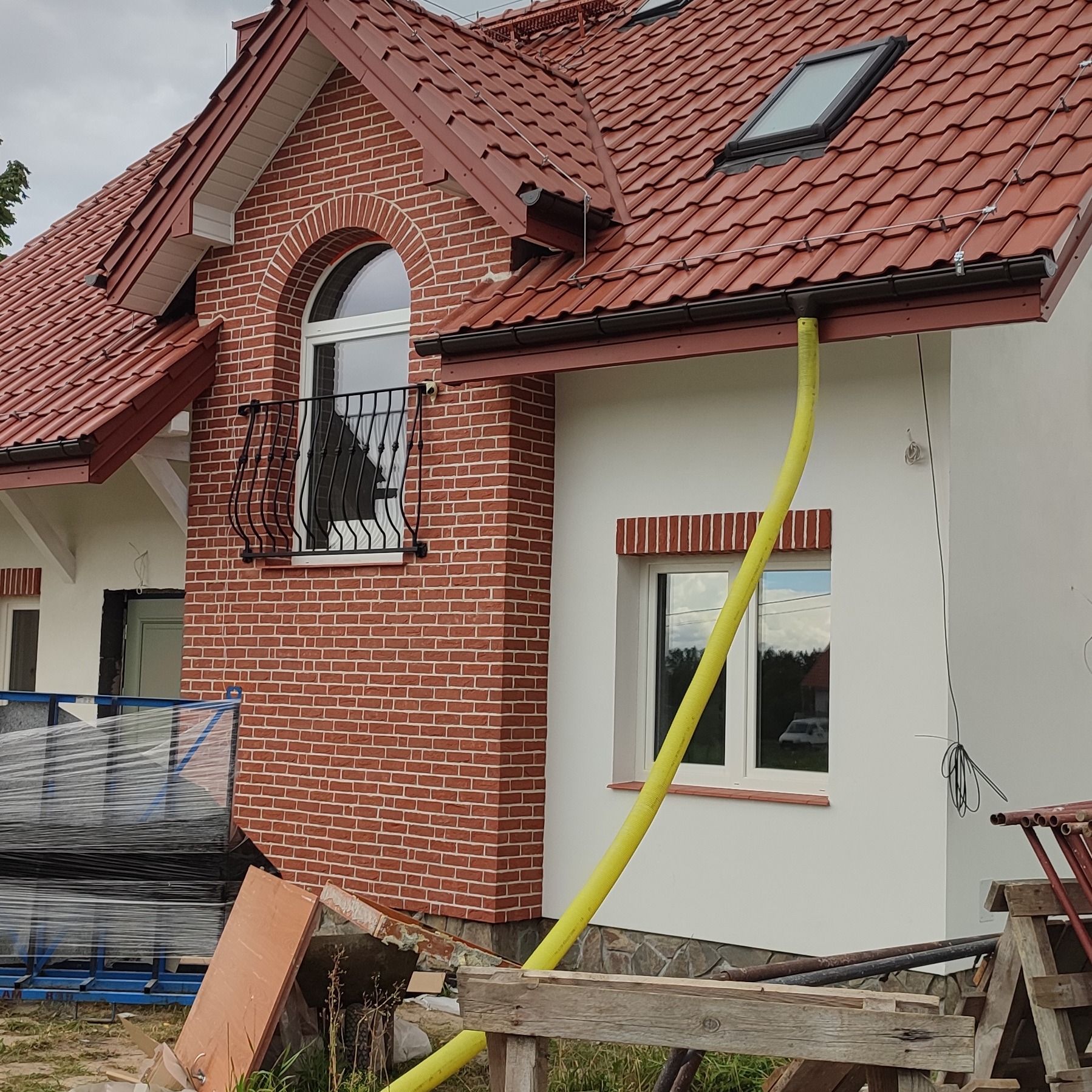 Dom jednorodzinny z czerwoną dachówką i elewacją w trakcie wykończenia, widoczna żółta rura wentylacyjna, rusztowania i materiały budowlane.