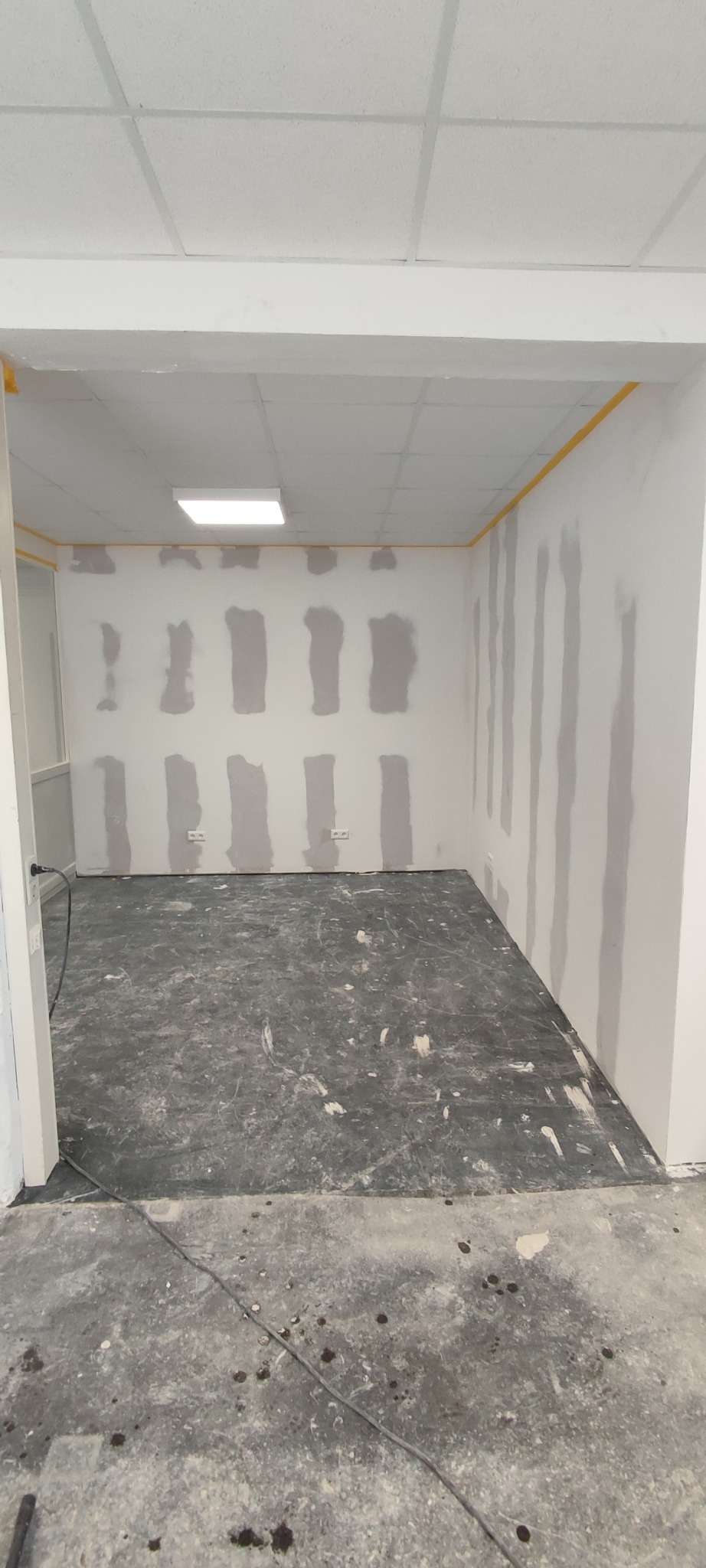 Pokój w trakcie szpachlowania ścian, zabezpieczony taśmą malarską przy suficie, z widocznymi śladami prac na podłodze i gładzią na ścianach.