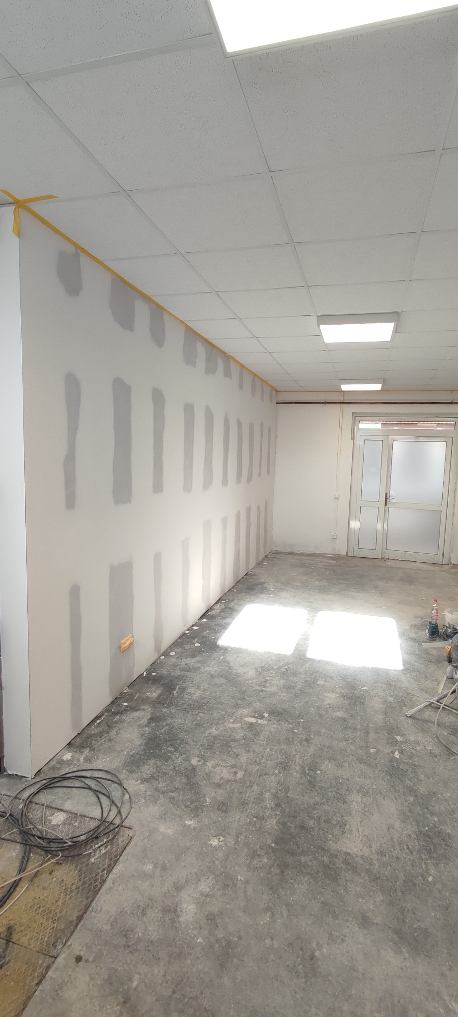 Wnętrze w trakcie szpachlowania: ściany z nałożonymi pasami gładzi, zabezpieczone taśmą, sufit kasetonowy, betonowa podłoga z plamami światła.