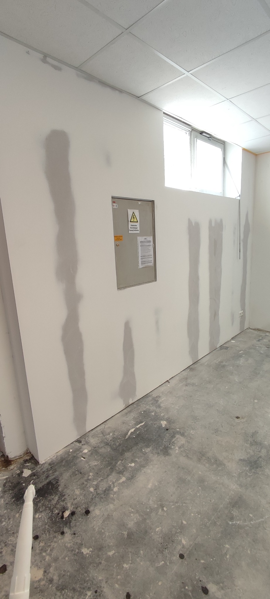 Ściana w trakcie szpachlowania z widocznymi pionowymi pasami świeżo nałożonej gładzi, skrzynka elektryczna na ścianie, surowa podłoga, widok z dołu.