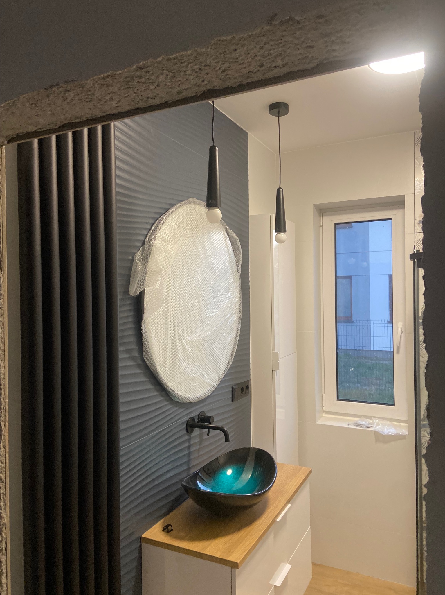 Nowoczesna łazienka w trakcie wykańczania: umywalka w odcieniach zieleni i czerni, okrągłe lustro w folii ochronnej, czarna armatura, dekoracyjna ściana z pionowymi panelami oraz dwie wiszące lampy...