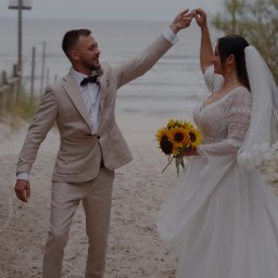Justyna Klimkowicz - Para młoda trzymająca się za ręce na piaszczystej plaży, panna młoda z bukietem słoneczników, pan młody w beżowym garniturze z muchą, drewniana ścieżka i morze w tle.