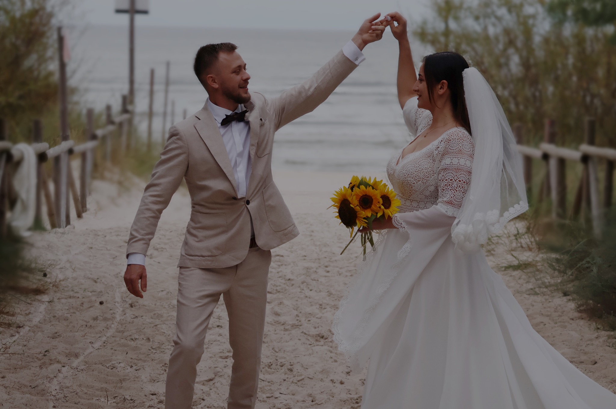 Para młoda trzymająca się za ręce na piaszczystej plaży, panna młoda z bukietem słoneczników, pan młody w beżowym garniturze z muchą, drewniana ścieżka i morze w tle.