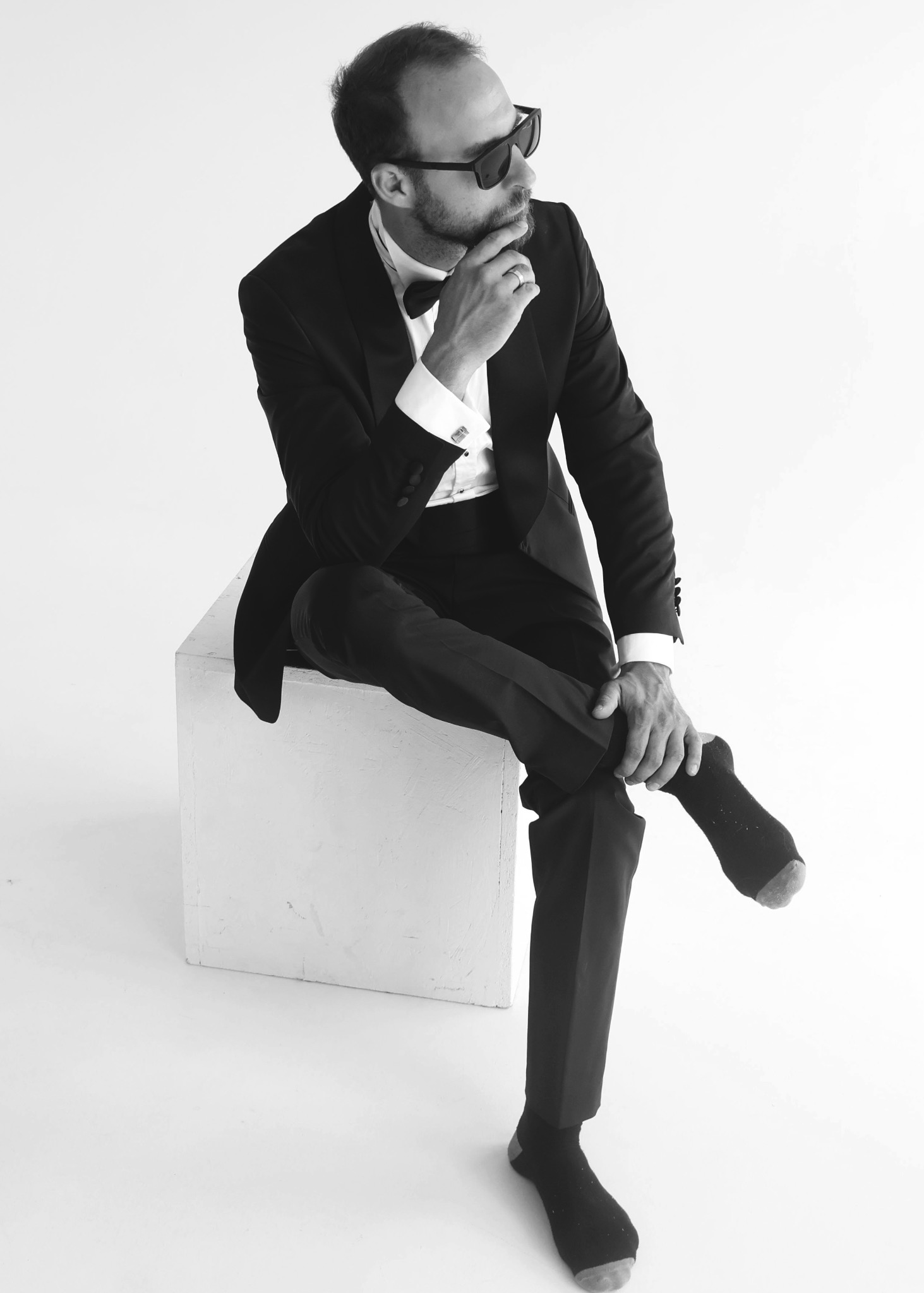Czarno-białe zdjęcie mężczyzny w smokingu i okularach przeciwsłonecznych, siedzącego na białym sześcianie, z jedną nogą w skarpetce trzymaną w dłoni, w zamyślonej pozie.