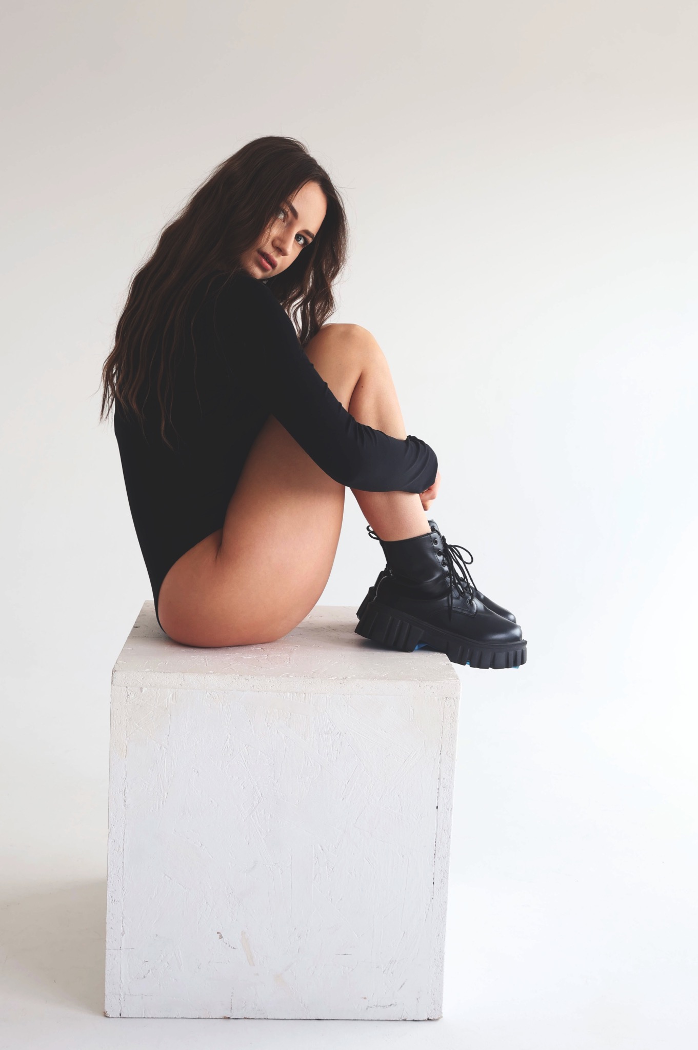 Kobieta w czarnym body i masywnych butach siedząca na białym kubiku na białym tle, patrząca w obiektyw.