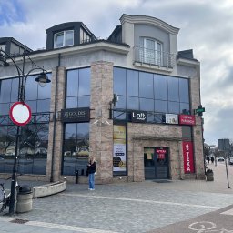 Goldis Nieruchomosci - Dwupiętrowy budynek z dużymi oknami, w których odbija się niebo, z szyldami agencji nieruchomości i apteki, stojący przy chodniku z kostki brukowej, z rowerem i znakiem drogowym na pierwszym planie.