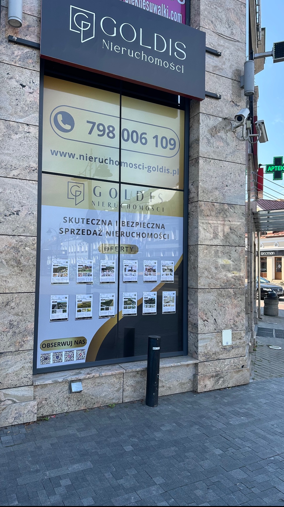 Fasada budynku z biurem nieruchomości Goldis, witryna z numerem telefonu, adresem strony internetowej i ofertami sprzedaży nieruchomości, widoczne kamery monitoringu.