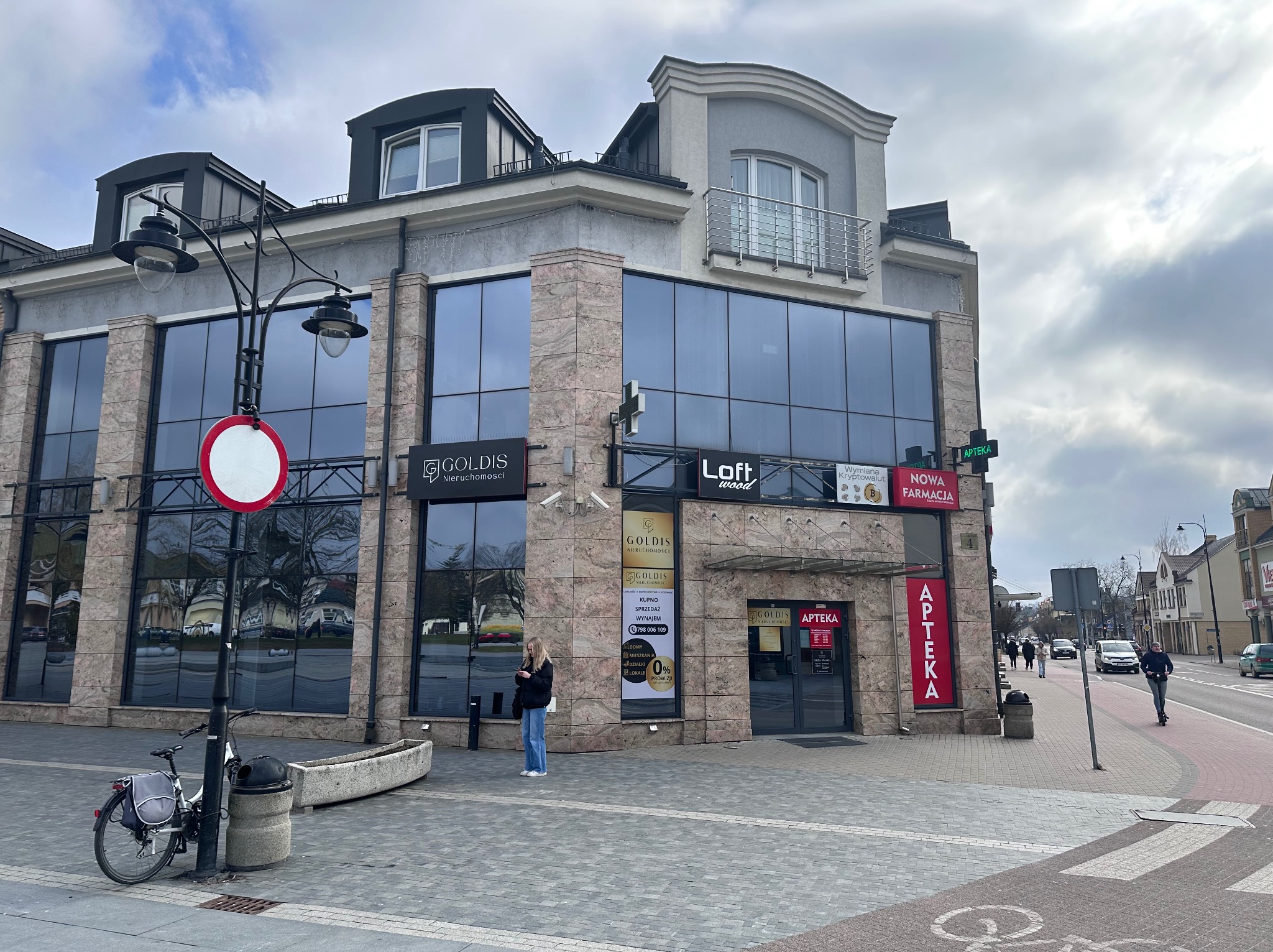 Dwupiętrowy budynek z dużymi oknami, w których odbija się niebo, z szyldami agencji nieruchomości i apteki, stojący przy chodniku z kostki brukowej, z rowerem i znakiem drogowym na pierwszym planie.
