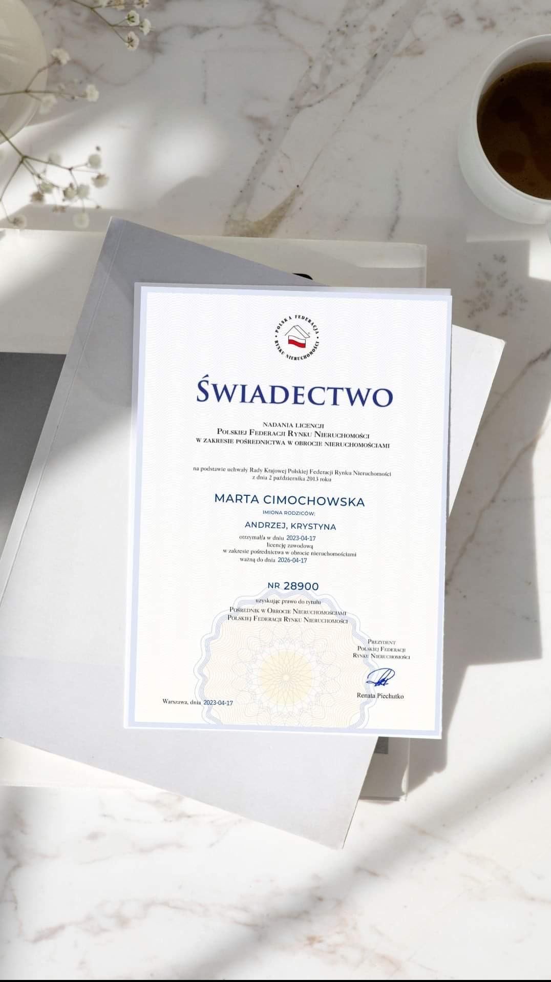 Świadectwo nadania licencji pośrednika w obrocie nieruchomościami, wydane przez Polską Federację Rynku Nieruchomości, z filigranem i podpisem, leżące na białym tle z dekoracyjnymi kwiatami...