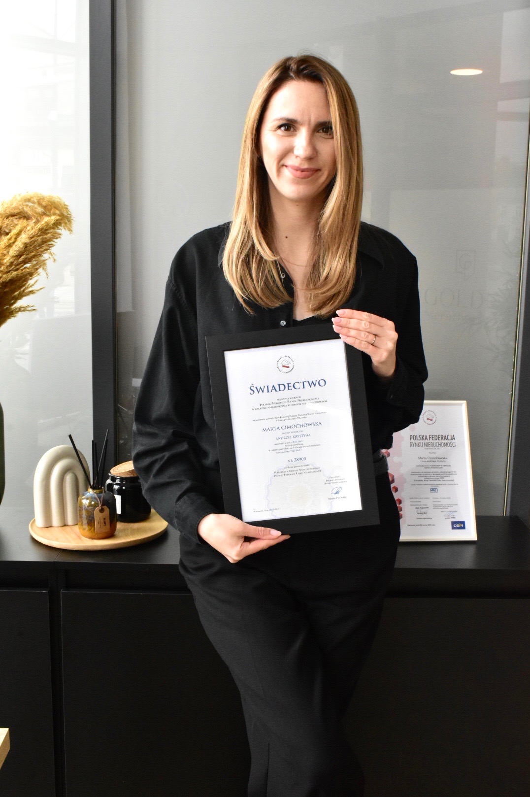 Kobieta w czarnej koszuli trzyma oprawione świadectwo licencji Polskiej Federacji Rynku Nieruchomości, na tle biurka z dekoracjami i drugim certyfikatem.
