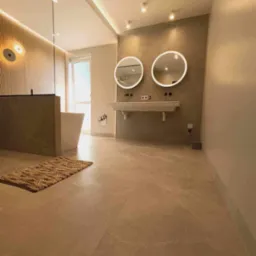 Nowoczesna łazienka z beżowymi płytkami podłogowymi, podświetlanymi okrągłymi lustrami nad podwieszaną umywalką i plecionym dywanikiem z juty.