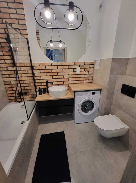 Nowoczesna łazienka z ceglaną ścianą, okrągłym lustrem z podwójnym oświetleniem, białą umywalką nablatową, czarną armaturą, wanną z parawanem i pralką pod blatem.