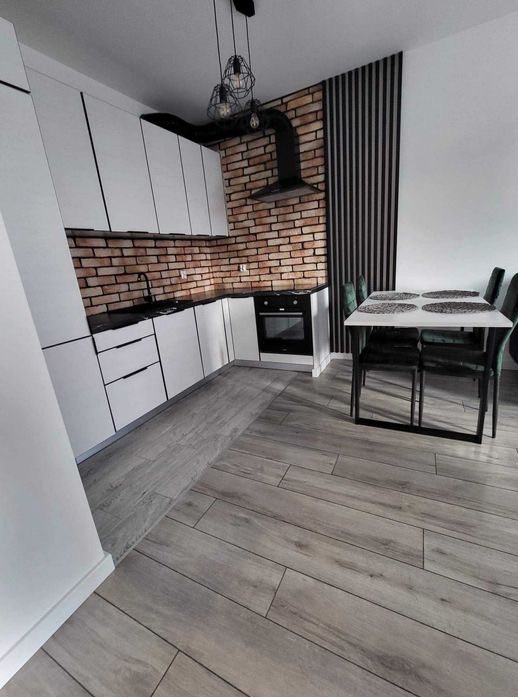Nowoczesna kuchnia z białymi szafkami, ceglaną ścianą i czarnym okapem, uzupełniona drewnopodobną podłogą i jadalnią z czarnymi krzesłami.