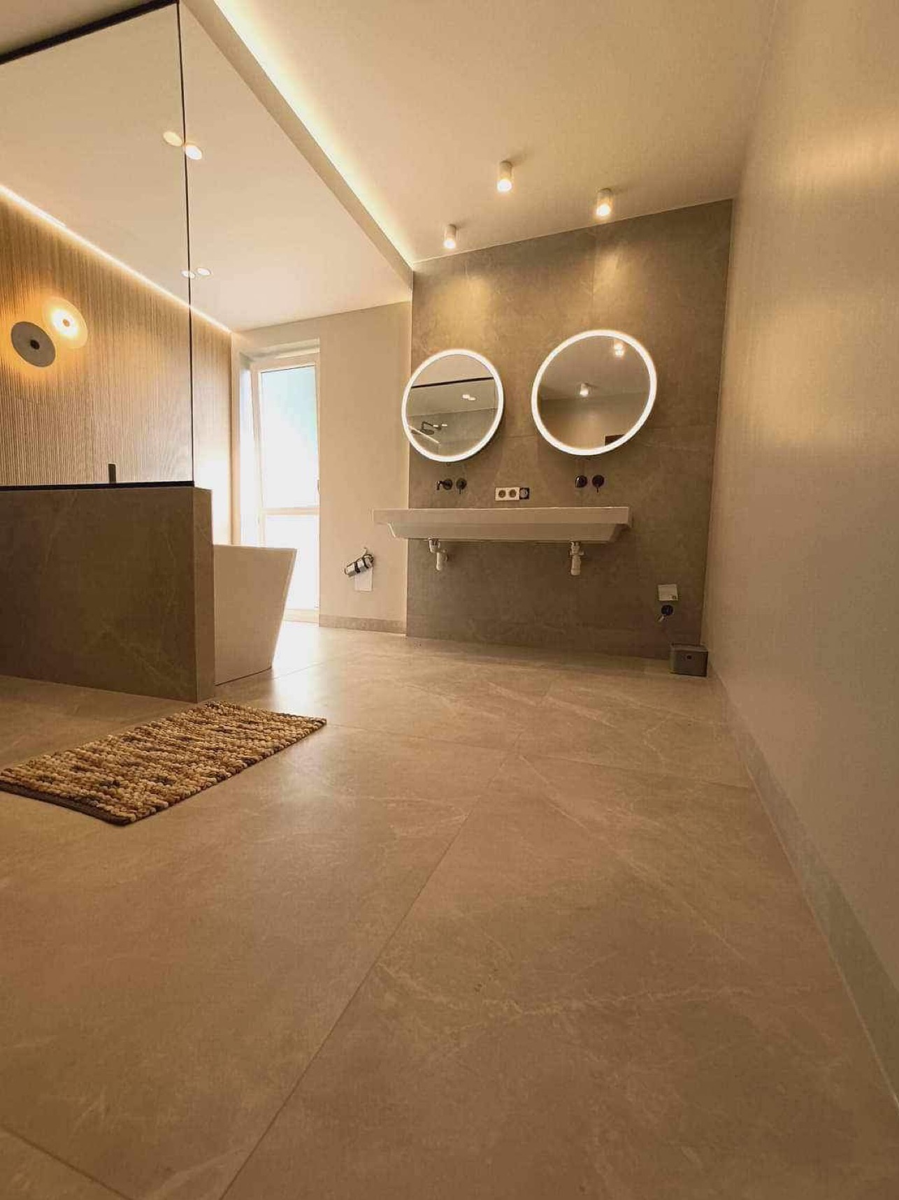 Nowoczesna łazienka z beżowymi płytkami podłogowymi, podświetlanymi okrągłymi lustrami nad podwieszaną umywalką i plecionym dywanikiem z juty.