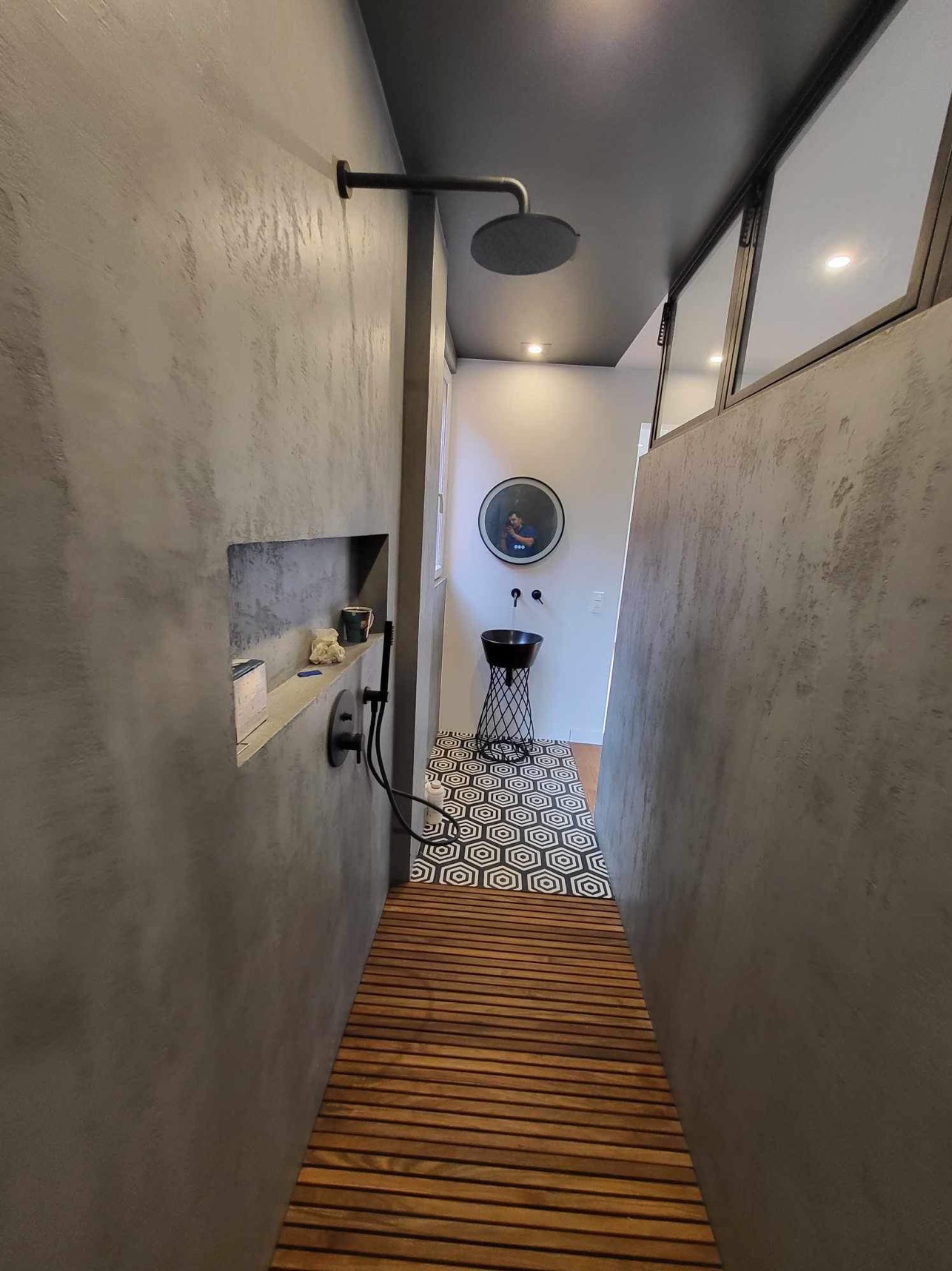 Nowoczesna łazienka z szarą, teksturowaną ścianą, drewnianą podłogą w strefie prysznica, czarną armaturą i umywalką na metalowym stelażu, heksagonalną płytką na podłodze oraz wnęką w ścianie...