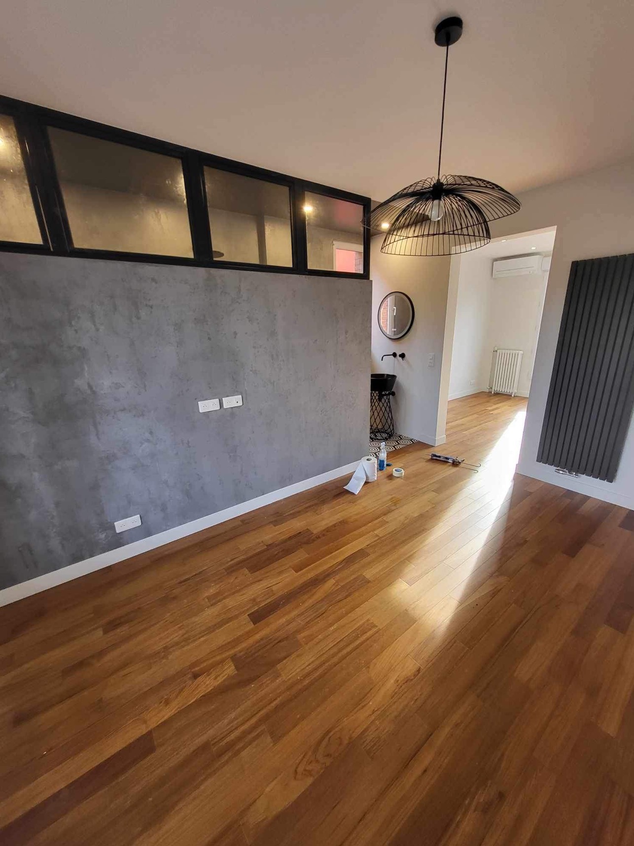 Wnętrze po remoncie z drewnianą podłogą, szarą ścianą z fakturą betonu i czarnymi akcentami: luksfery, lampa, umywalka i grzejnik.