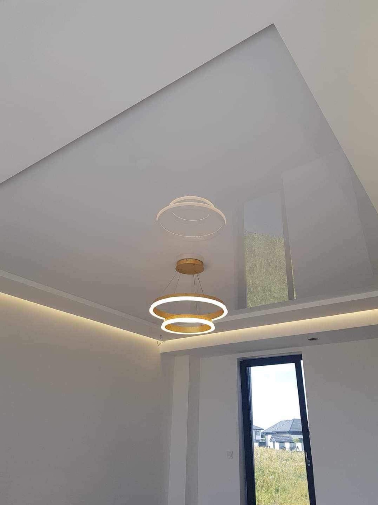 Biały sufit napinany z geometrycznym podziałem na płaszczyzny, oświetlony liniowymi LED-ami w listwach i nowoczesną, dwuokręgową lampą wiszącą z podświetleniem LED, odbijający się w lustrzanej...