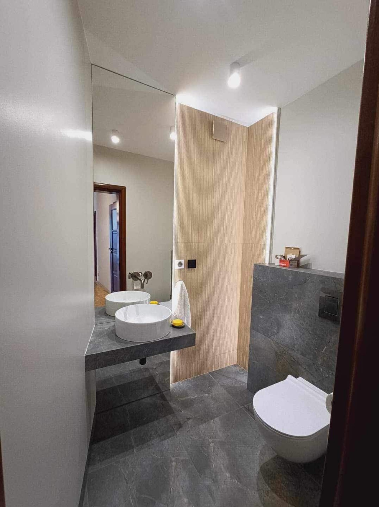 Nowoczesna łazienka z umywalką nablatową na szarym blacie, toaletą wiszącą na szarej ścianie z płytek imitujących kamień, lustrem na ścianie i drewnianą ścianą z pionowymi paskami oraz oświetleniem...