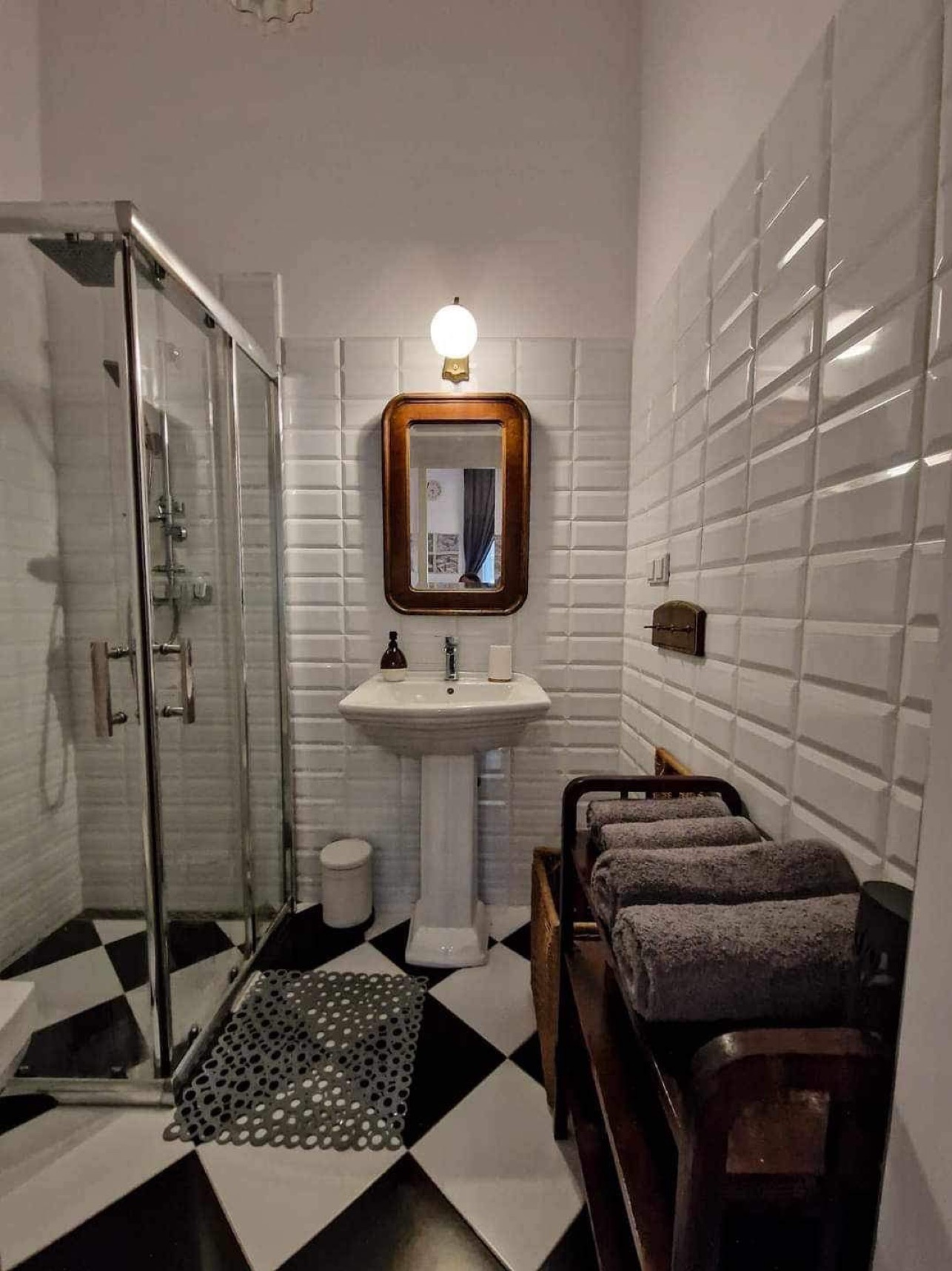 Biała łazienka w stylu retro z kabiną prysznicową, umywalką na postumencie, lustrem w drewnianej ramie, białą wypukłą glazurą, czarno-białą szachownicą na podłodze i stertą szarych ręczników...
