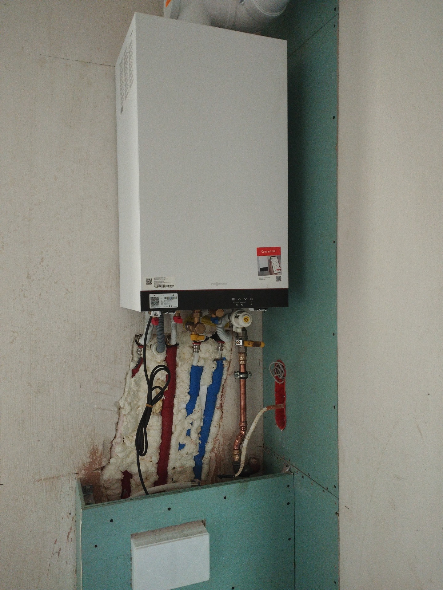 Nowy piec gazowy Viessmann zamontowany na ścianie z płyt kartonowo-gipsowych, z widocznymi rurami i przewodami izolowanymi pianką montażową w kolorach czerwonym i niebieskim.