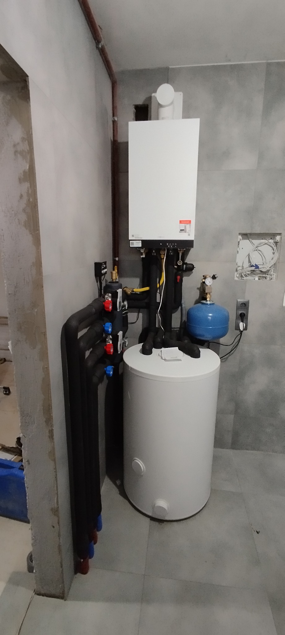 Biały piec gazowy kondensacyjny z podłączonym zasobnikiem ciepłej wody, rurami w izolacji i niebieskim naczyniem przeponowym, zamontowany na szarej płytce w pomieszczeniu w trakcie wykańczania.