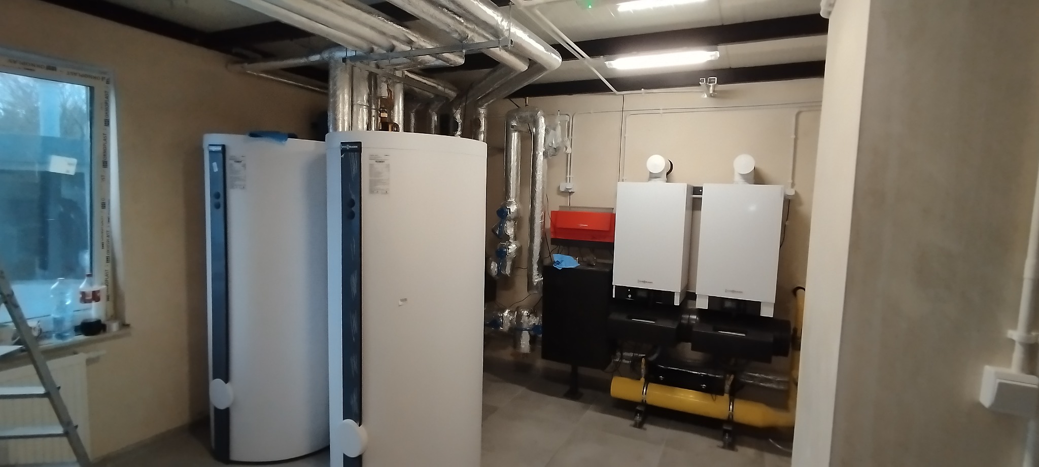 Dwa białe zbiorniki buforowe Viessmann obok okna, z dużą ilością rur instalacyjnych pod sufitem i dwoma piecami grzewczymi Viessmann na czarnym stelażu z żółtym elementem, w pomieszczeniu technicznym.