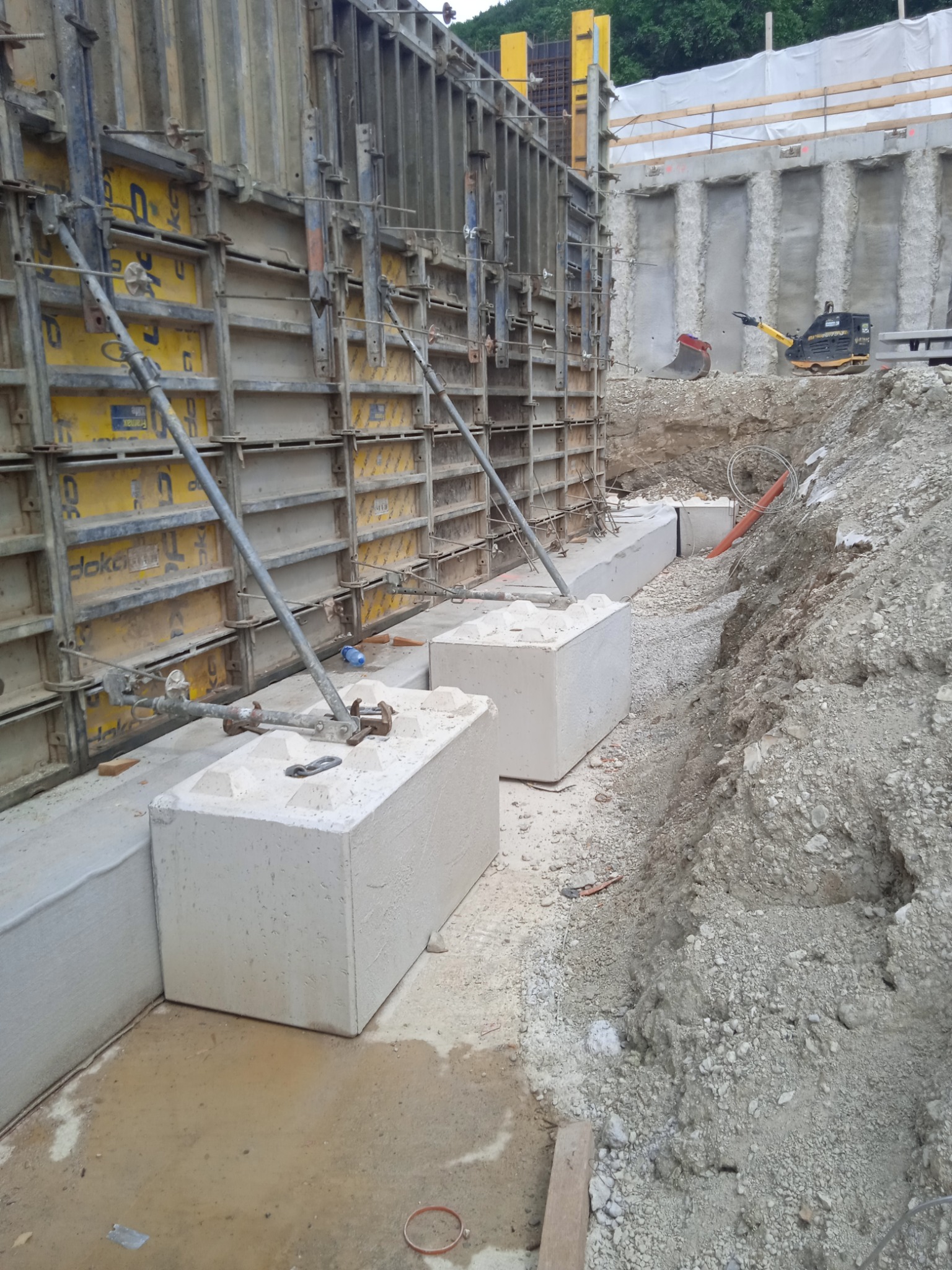Szczegółowe ujęcie szalunków fundamentowych z żółtymi panelami i betonowych bloków oporowych na placu budowy, z ubijarką w tle.
