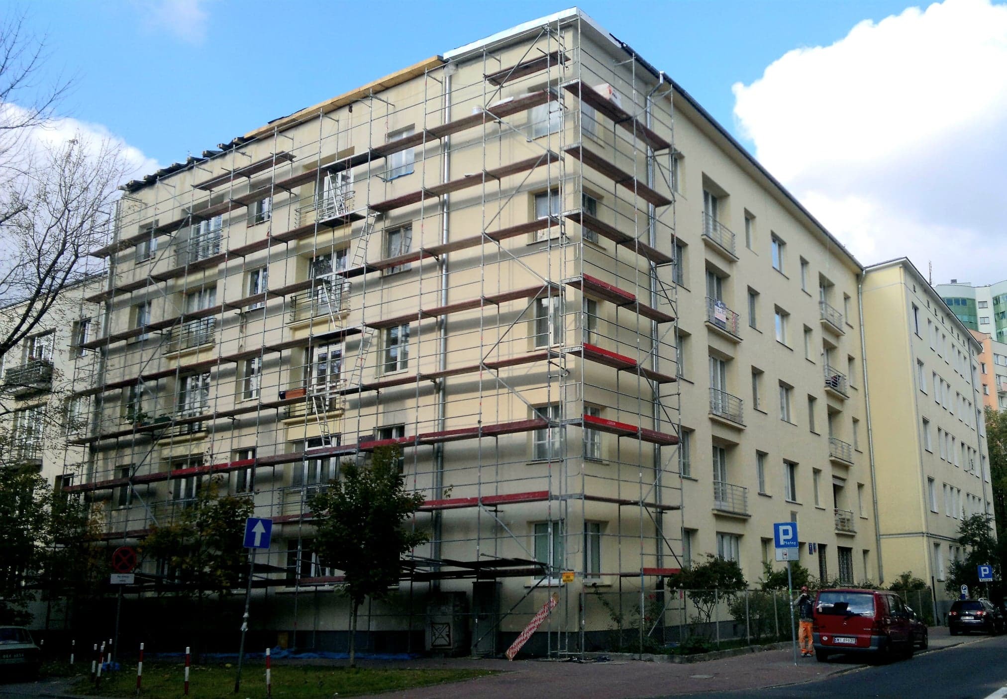 Wielopiętrowy budynek mieszkalny w trakcie prac elewacyjnych, osłonięty rusztowaniami, widoczny fragment chodnika z zaparkowanymi samochodami i znakami drogowymi.