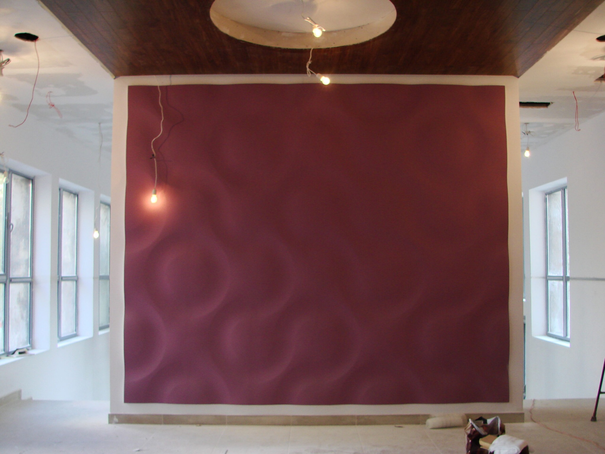 Ściana pomalowana na bordowo z subtelnym wzorem falistym, widoczne okna z białymi ramami i fragment sufitu z drewnianymi panelami, surowy stan wykończenia wnętrza z widocznymi kablami i narzędziami.