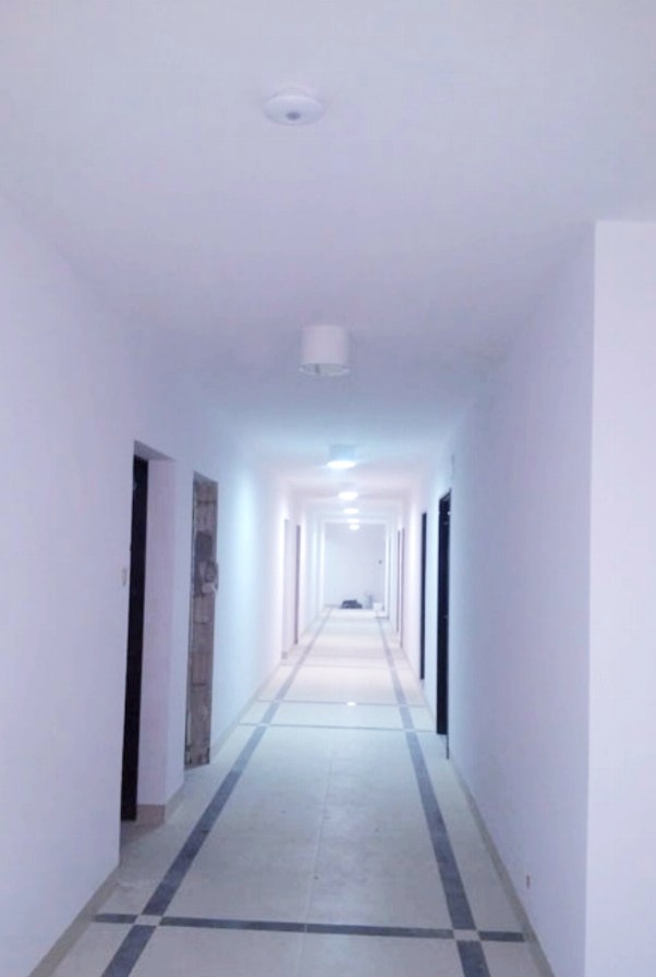Długi korytarz w budynku po świeżym malowaniu ścian na biało, z szarymi płytkami podłogowymi obramowanymi ciemnymi pasami i prostymi lampami sufitowymi.