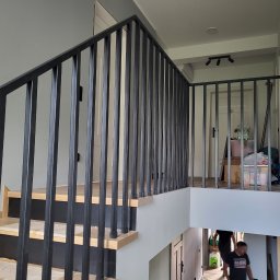 Loft Antima - Nowoczesne schody z drewnianymi stopniami i czarną metalową balustradą z pionowych prętów, widok z dołu na górę, w tle jasne ściany i drzwi.