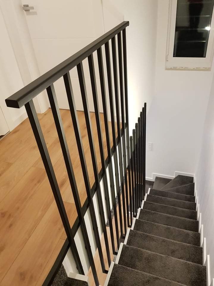 Nowoczesna, czarna balustrada z pionowych prętów zamontowana przy schodach z szarą wykładziną, widok z góry na dół.