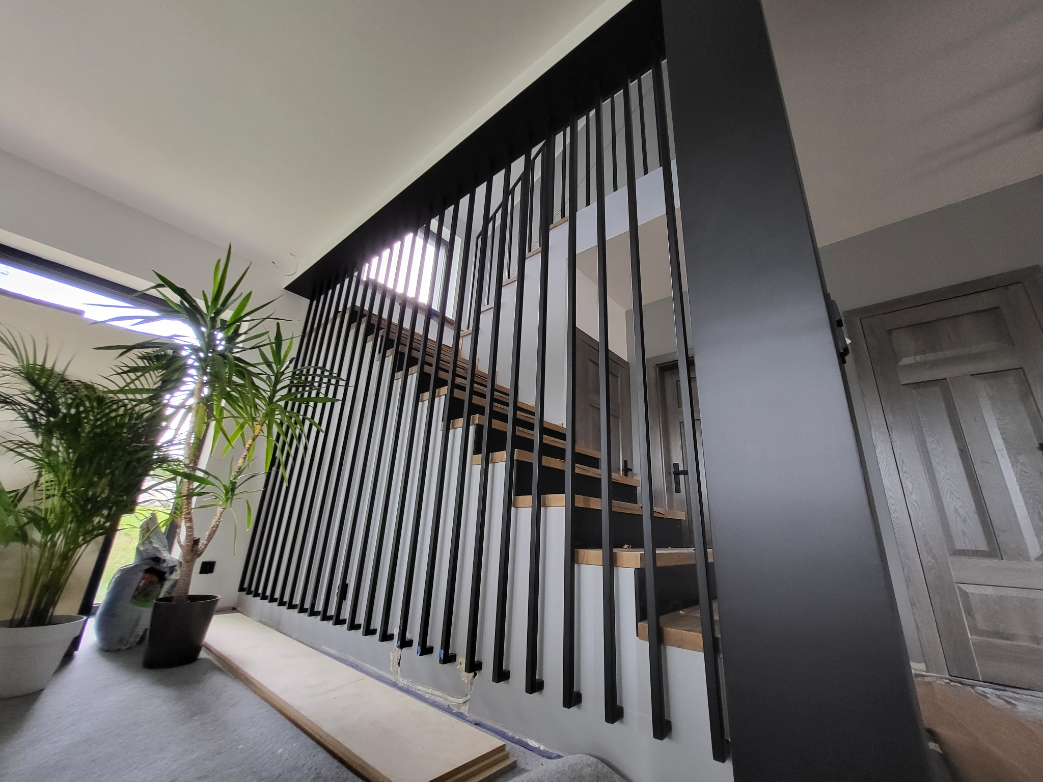 Nowoczesne schody z drewnianymi stopniami i czarną, minimalistyczną balustradą z pionowych prętów, widoczne rośliny doniczkowe i materiały budowlane wskazują na trwający montaż.