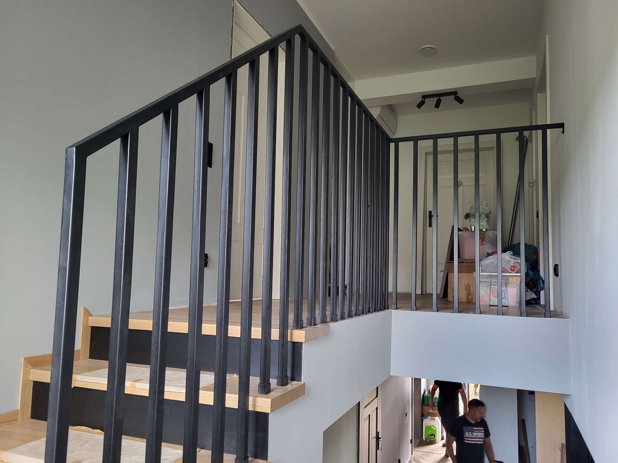 Nowoczesne schody z drewnianymi stopniami i czarną metalową balustradą z pionowych prętów, widok z dołu na górę, w tle jasne ściany i drzwi.