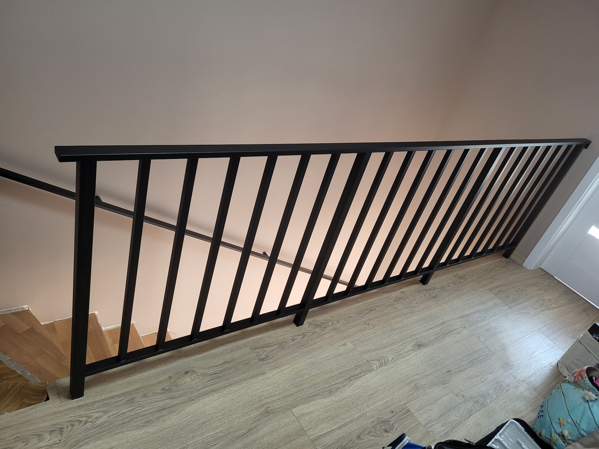 Czarna, metalowa balustrada schodowa z pionowymi szczeblami, widok z góry na drewniane schody i podłogę imitującą drewno.