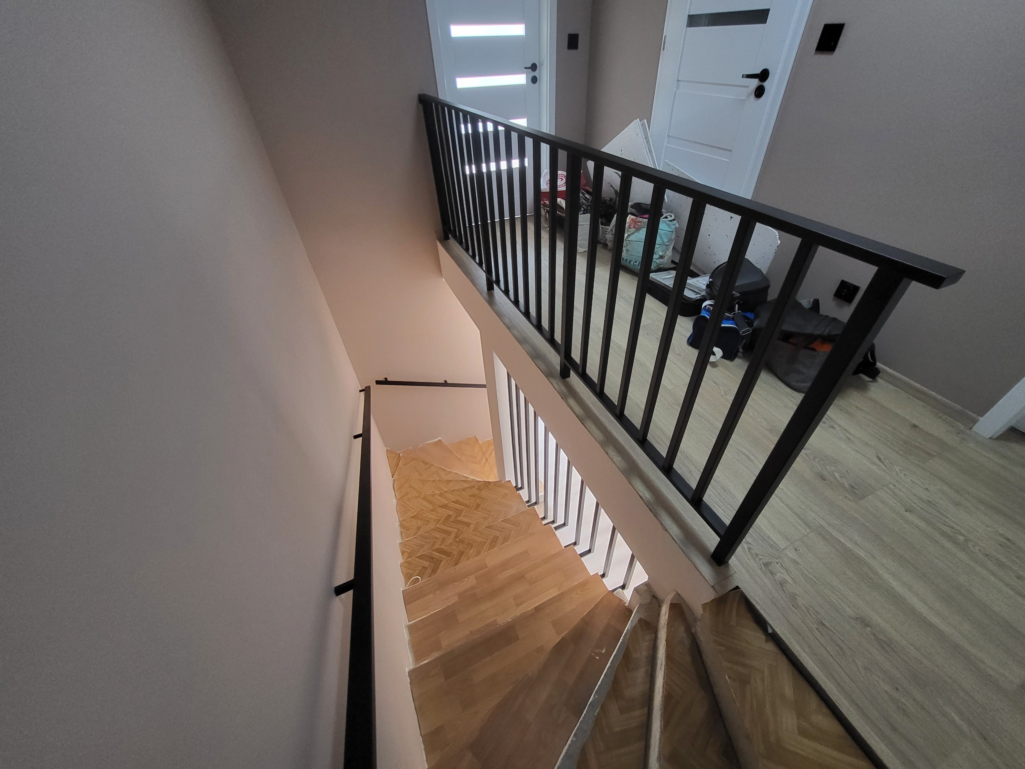 Widok z góry na drewniane schody z parkietem w jodełkę, zabezpieczone czarną, metalową balustradą z pionowymi szczebelkami. W tle widoczne białe drzwi wewnętrzne.