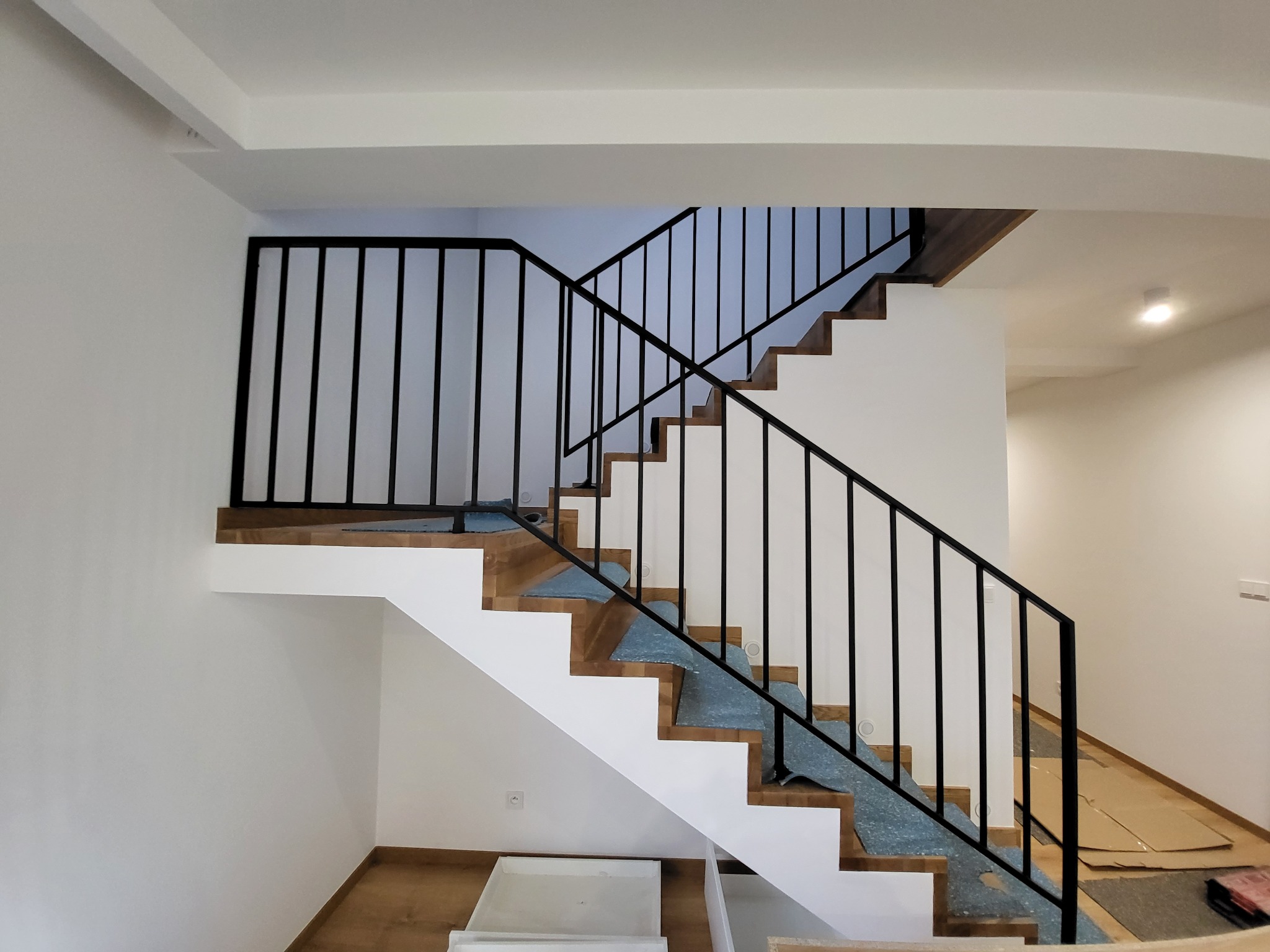 Nowoczesne schody z drewnianymi stopniami, białymi podstopnicami i czarną metalową balustradą, zabezpieczone folią ochronną podczas prac wykończeniowych.