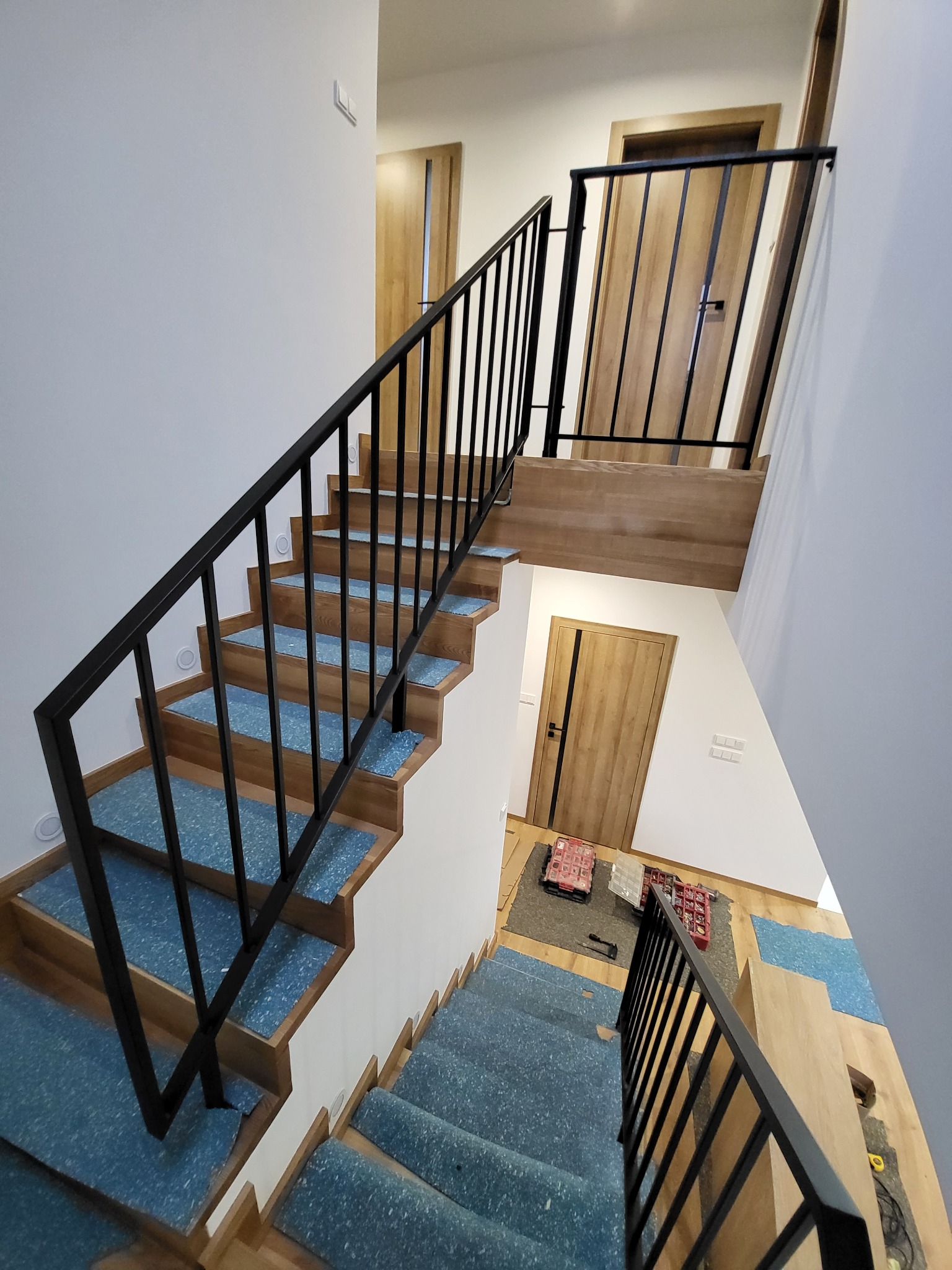 Wnętrze domu z drewnianymi schodami zabezpieczonymi folią, czarne metalowe poręcze, widok z góry na dwa poziomy z drzwiami.