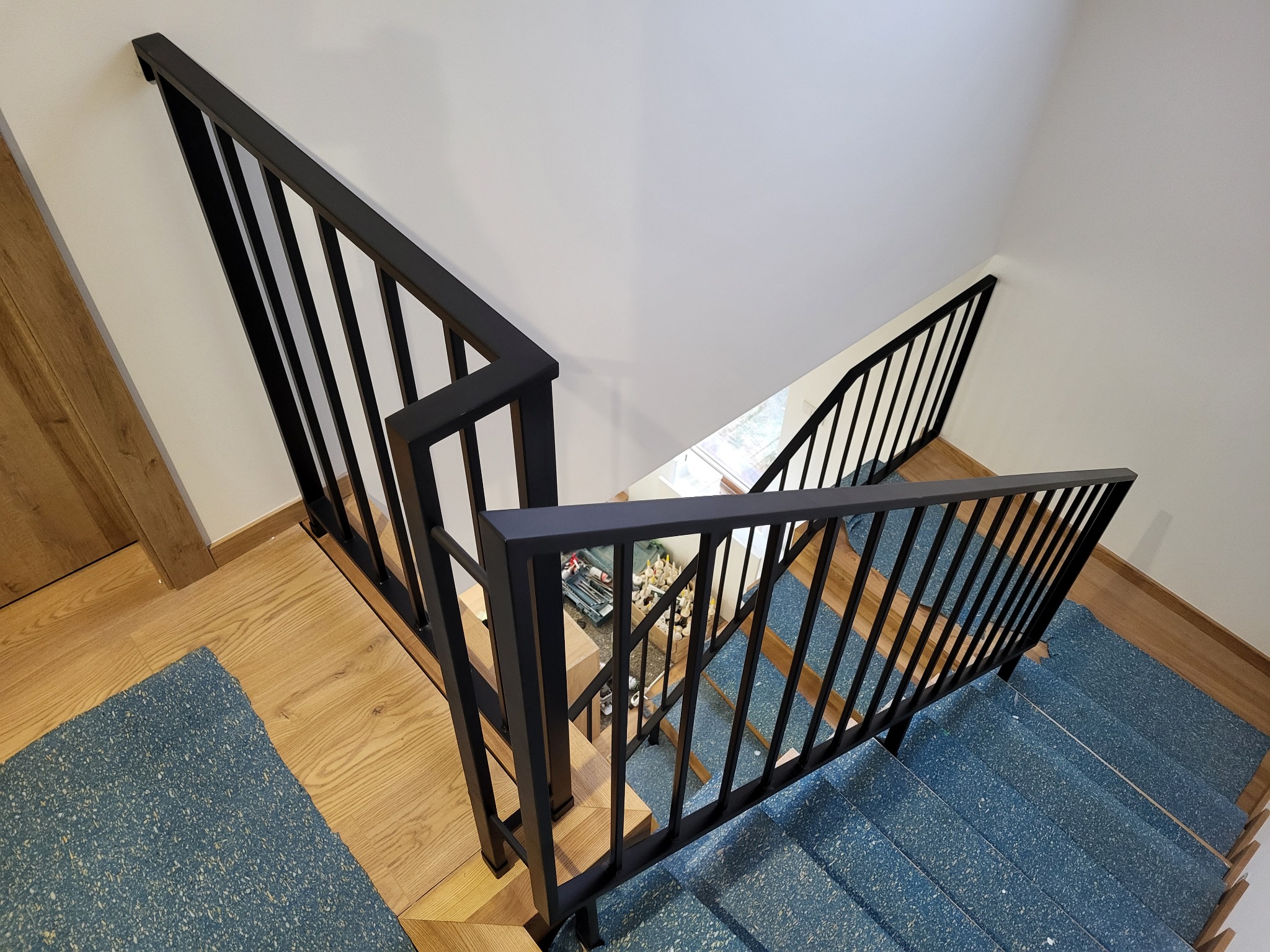 Widok z góry na schody z drewnianymi stopniami pokrytymi niebieską wykładziną i czarną metalową balustradą, skrzynka narzędziowa widoczna na podeście.