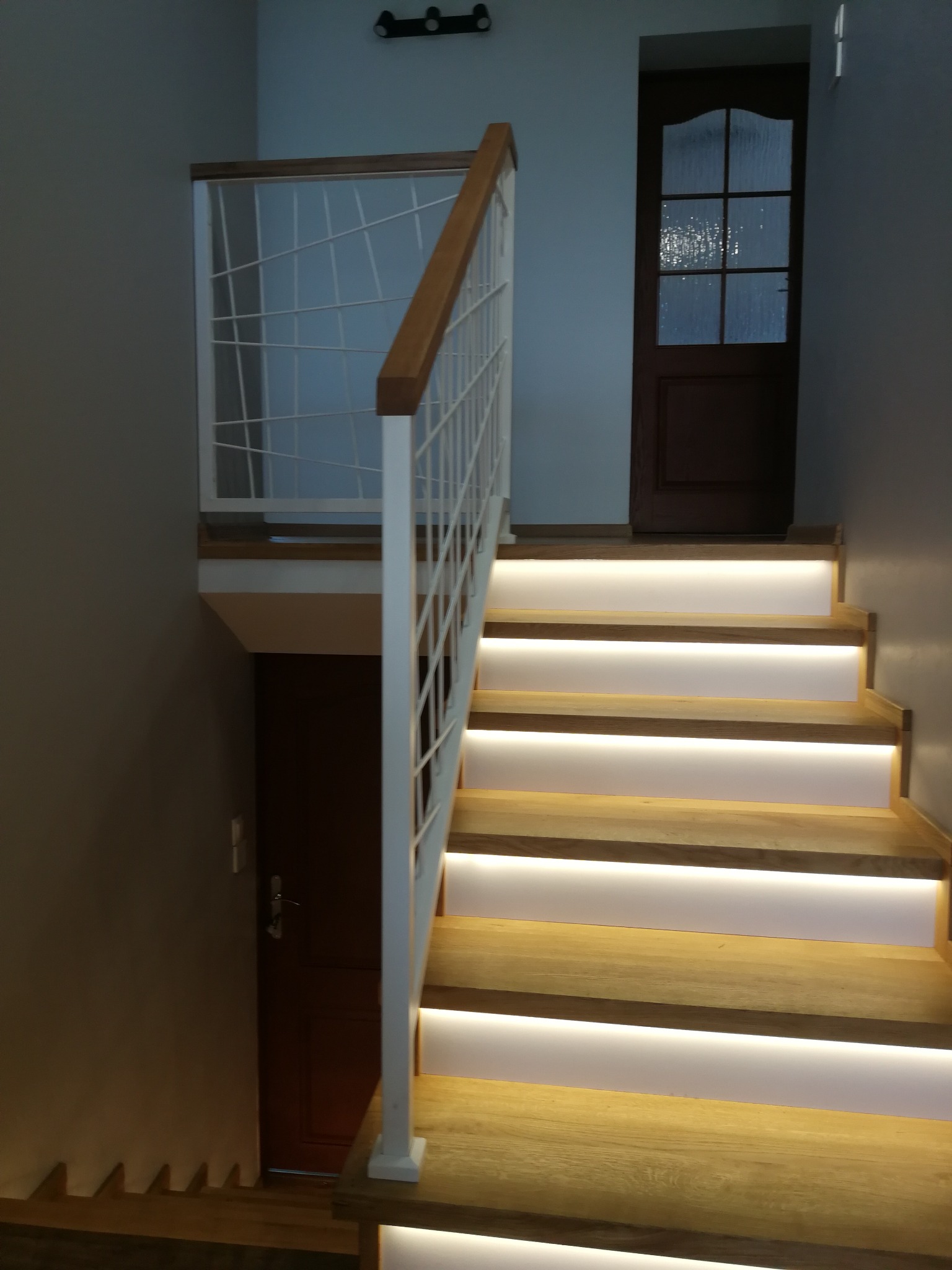 Nowoczesne schody drewniane z podświetleniem LED w Kostrzynie, z białą balustradą z metalowymi prętami i drewnianą poręczą, prowadzące do ciemnych drzwi.