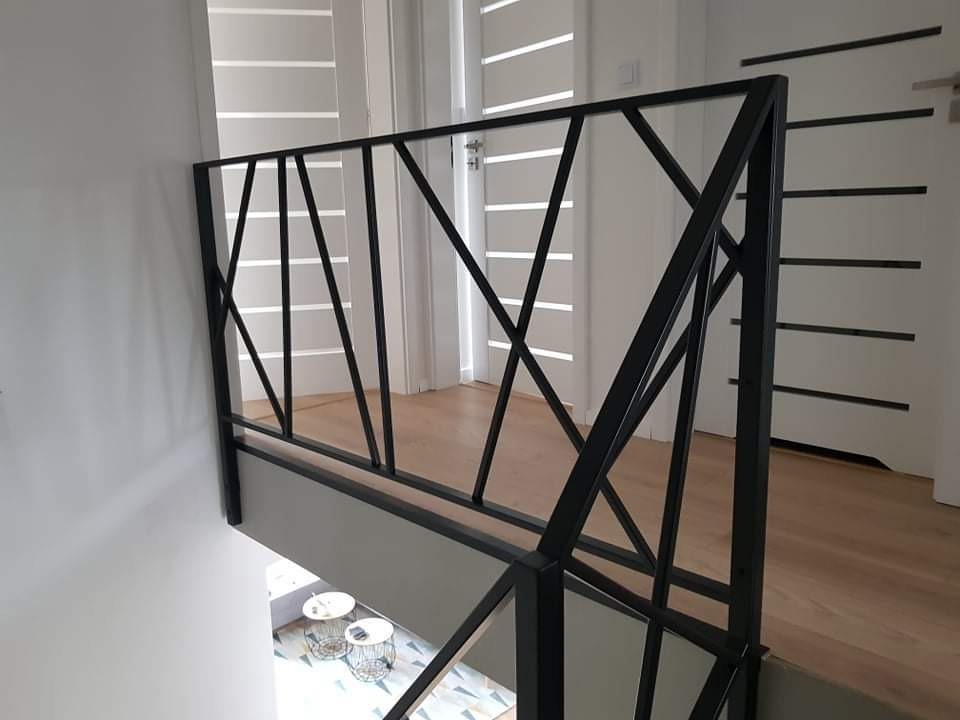Czarna, metalowa balustrada schodowa o geometrycznym wzorze w nowoczesnym wnętrzu z drewnianą podłogą i białymi drzwiami z poziomymi zdobieniami.