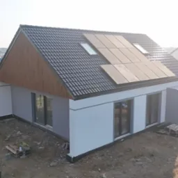 Nowoczesny, biało-szary dom z panelami słonecznymi na dachu, widok z góry na plac budowy z paletami i wężem.