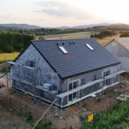 Budowa domu jednorodzinnego z czarną dachówką i oknami dachowymi, w tle krajobraz z górami i zachodzącym słońcem, widoczne rusztowania.