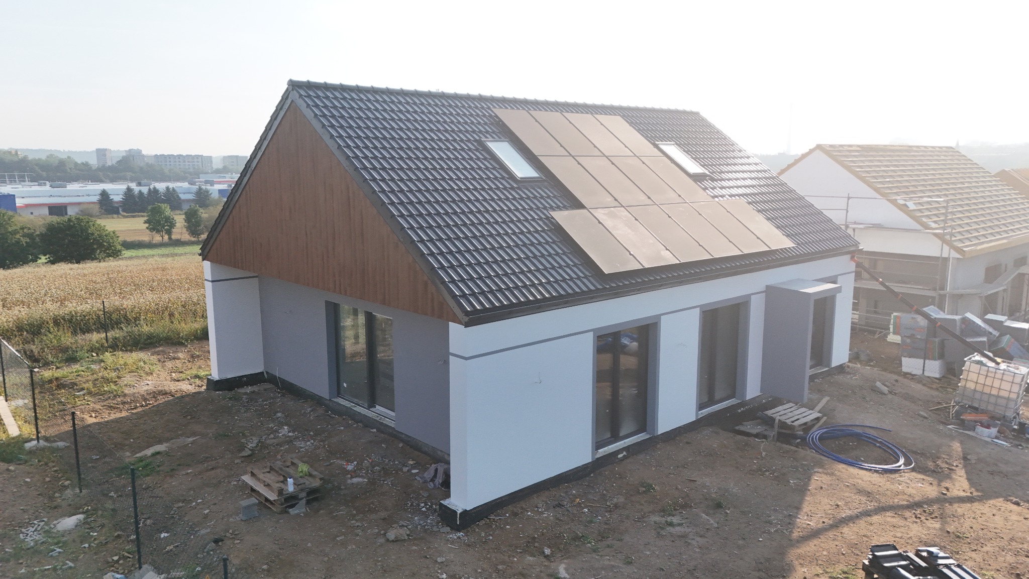 Nowoczesny, biało-szary dom z panelami słonecznymi na dachu, widok z góry na plac budowy z paletami i wężem.