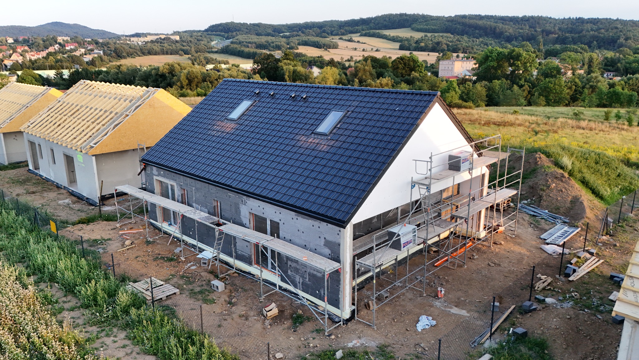 Wysoki kąt widzenia na plac budowy z trzema domami w różnym stanie ukończenia, jeden z czarną dachówką i ociepleniem ścian zewnętrznych, rusztowania dookoła, w tle zielone wzgórza i zabudowania.