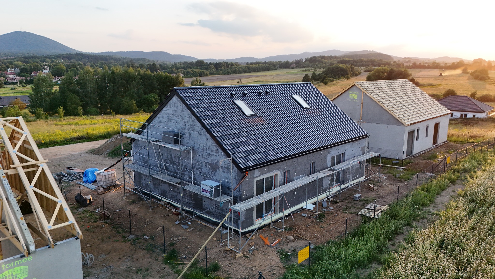Budowa domu jednorodzinnego z czarną dachówką i oknami dachowymi, w tle krajobraz z górami i zachodzącym słońcem, widoczne rusztowania.