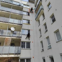 Pro-Alpin - Dwóch alpinistów na linach, zabezpieczających okna taśmą, pracuje na elewacji wysokiego bloku z balkonami. Widok z dołu, błękitne niebo w tle.