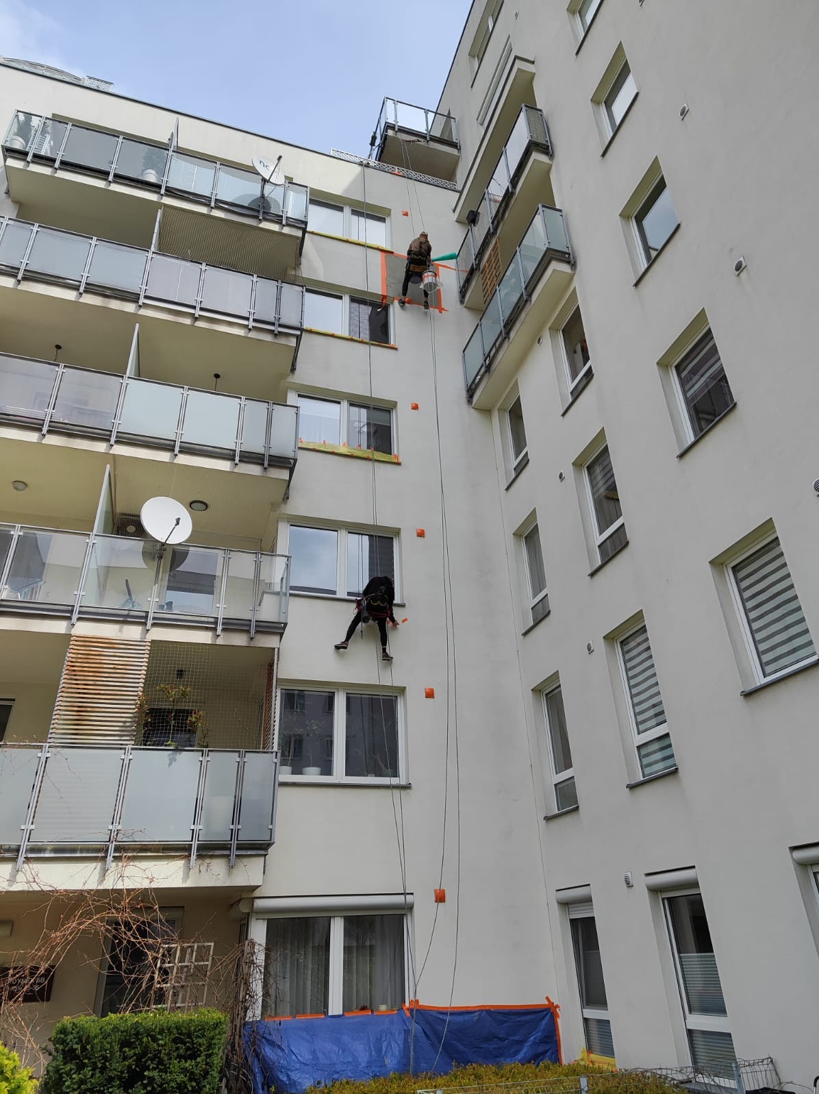 Dwóch alpinistów na linach, zabezpieczających okna taśmą, pracuje na elewacji wysokiego bloku z balkonami. Widok z dołu, błękitne niebo w tle.