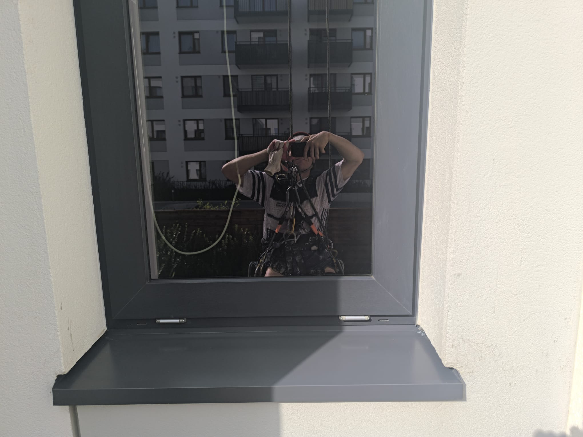 Szare okno z parapetem. W szybie odbicie osoby w uprzęży, robiącej zdjęcie. W tle budynek mieszkalny. Widoczne liny i elementy zabezpieczające.