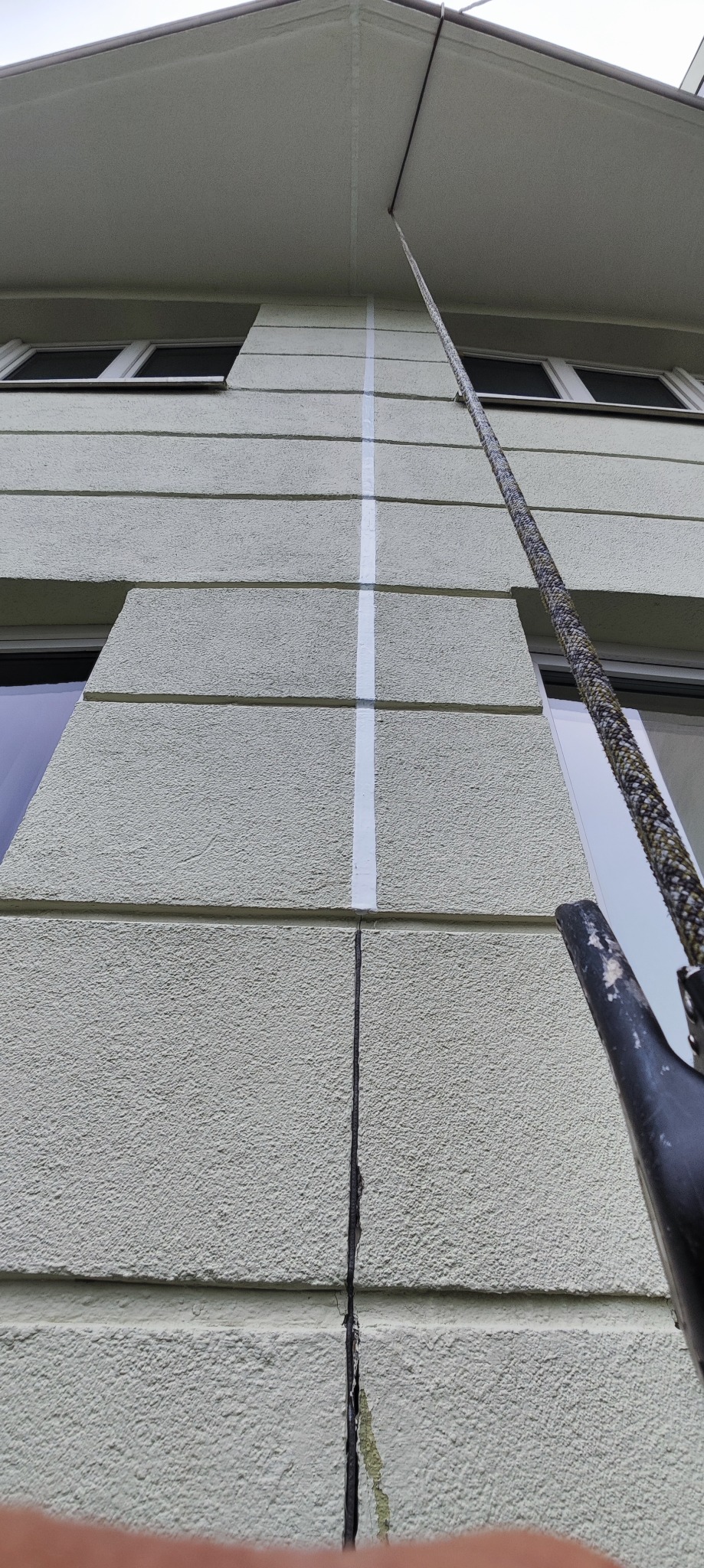 Jasna elewacja budynku z widoczną dylatacją pionową, zabezpieczona taśmą malarską. Ujęcie z dołu, widoczny fragment liny i okna.